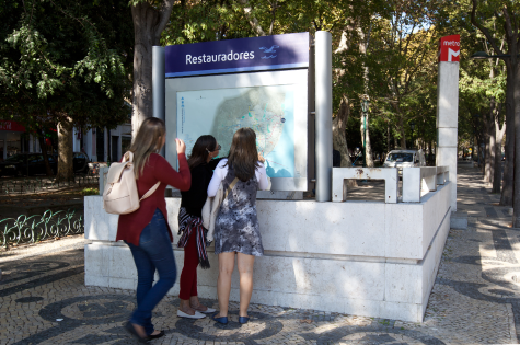 Fotografia com três raparigas jovens junto a um acesso do Metro, olhando para um mapa da cidade e Metro.